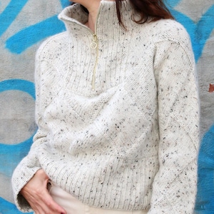 Puede incluir: Un suéter de punto crema moteado con cuello alto y cremallera dorada. El suéter tiene un patrón texturizado y mangas largas. La modelo lleva pantalones color crema.