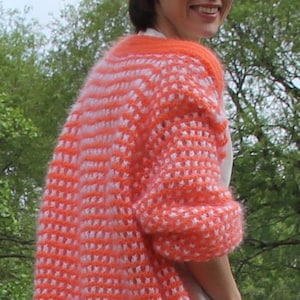 Könnte beinhalten: Ein offener, orange-weiß gehäkelter Cardigan. Der langärmlige Pullover hat ein strukturiertes Muster und einen durchgehenden orangefarbenen Rand. Der Cardigan wird über einem weißen Oberteil und einer blauen Jeans getragen. Der Hintergrund umfasst eine weiße Brücke und grüne Bäume.