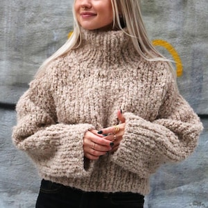 Peut inclure: Un pull à col roulé beige en tricot épais avec des manches larges. Le pull court est fait d'un fil épais et texturé. Le modèle porte un jean noir. Le pull est de couleur neutre, adapté à l'automne et à l'hiver.