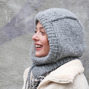 Könnte beinhalten: Eine graue Strick-Balaclava mit passendem Schal. Die Balaclava bedeckt Kopf und Hals, mit einer Gesichtsöffnung. Die Person trägt eine cremefarbene, flauschige Jacke.