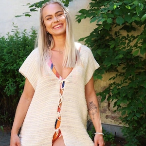 Könnte beinhalten: Ein cremefarbenes, kurzärmeliges Häkeltop mit Schnürung vorne. Das Top ist offen und zeigt einen bunten Badeanzug darunter. Die Person lächelt, hat lange blonde Haare und steht vor grünem Laub.
