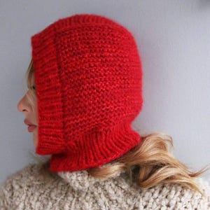 May include: A bright red knitted balaclava. The hood, made from a thick, textured yarn, covers the head and neck. The balaclava is a solid colour, with a visible knit pattern. The person is wearing a beige sweater.