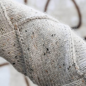 K&ouml;nnte beinhalten: Nahaufnahme eines hellgrauen Strickstoffs mit strukturierter Optik. Das Strickwerk weist ein Zopfmuster und Flecken dunklerer Farben auf. Der Stoff wirkt weich und warm, geeignet f&uuml;r einen Pullover oder eine Decke.