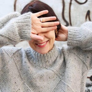 K&ouml;nnte beinhalten: Ein hellgrauer, grob gestrickter Pullover mit hohem Kragen und Rautenmuster. Der Pullover hat Farbflecken. Die Person l&auml;chelt und bedeckt ihre Augen mit der Hand.