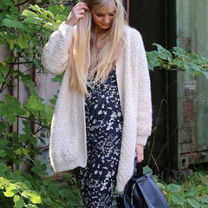 May include: A woman wearing a cream-coloured, open-front cardigan over a dark blue floral print dress. She is holding a black leather backpack. The background includes green foliage and a weathered metal structure.