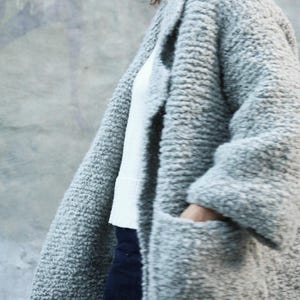 Peut inclure: Un manteau ouvert gris clair à la texture bouclée, doté d'un tricot épais et granuleux. Ce vêtement présente des manches longues, un large revers et deux grandes poches plaquées sur le devant, porté sur un haut blanc et un pantalon bleu.
