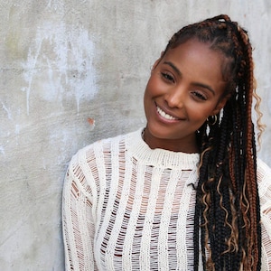 May include: A woman with dark skin and long, braided hair smiles at the camera. She wears a cream-colored, open-knit sweater. The background is a light gray wall.