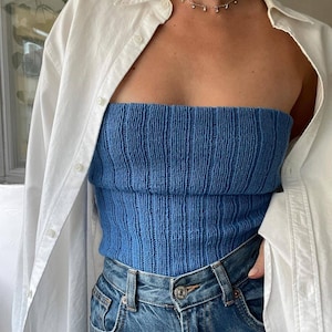 Może przedstawiać: Niebieski dzianinowy top z prążkowaną fakturą połączony z niebieskimi dżinsami. Biała koszula zapinana na guziki jest noszona rozpięta. Widoczny jest delikatny srebrny naszyjnik z małymi zawieszkami.