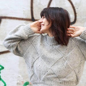 K&ouml;nnte beinhalten: Ein hellgrauer, strukturierter Strickpullover mit Rautenmuster. Der Pullover hat einen hohen, gerippten Kragen und gerippte B&uuml;ndchen. Die Person auf dem Bild l&auml;chelt und tr&auml;gt eine schwarze Hose.