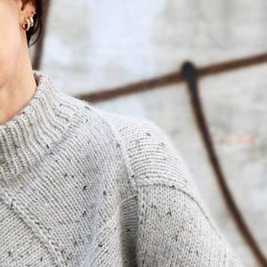 K&ouml;nnte beinhalten: Ein hellgrauer Strickpullover mit geripptem Rundhalsausschnitt und gesprenkelten Details. Der Pullover hat ein strukturiertes Aussehen und einen klassischen, bequemen Stil. Schulter und &Auml;rmel sind sichtbar.