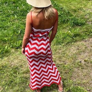 Puede incluir: Un vestido largo de crochet con estampado de chevrón rojo y blanco con finas correas rojas. El vestido se combina con un sombrero de sol de paja. La persona está de pie en una zona de césped.