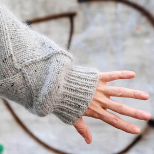 K&ouml;nnte beinhalten: Nahaufnahme eines hellgrauen Strickpullover&auml;rmels. Der &Auml;rmel hat ein geripptes B&uuml;ndchen und ein strukturiertes Strickmuster. Der Pullover scheint handgefertigt zu sein, mit sichtbaren Stichdetails und einem gesprenkelten Garn.