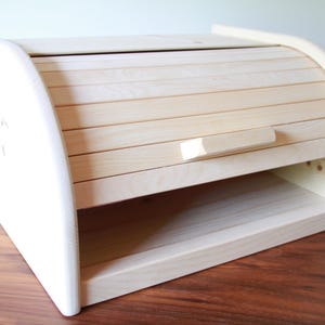 Könnte beinhalten: Ein helles Brotbox aus Holz mit einem Rolldeckel und einem kleinen Griff. Die Box hat eine rechteckige Basis mit einem Regal und gebogenen Seiten. Die Holzmaserung ist auf dem Deckel und der Vorderseite sichtbar.