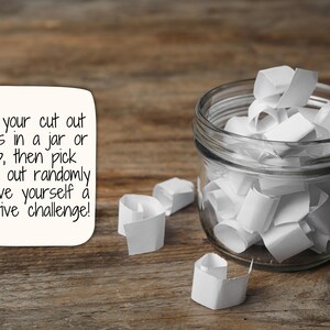 May include: A glass jar filled with white paper slips, with a few slips scattered on a wooden surface. The text on the white card reads: "Put your cut out cards in a jar or cup, then pick them out randomly to give yourself a creative challenge!"