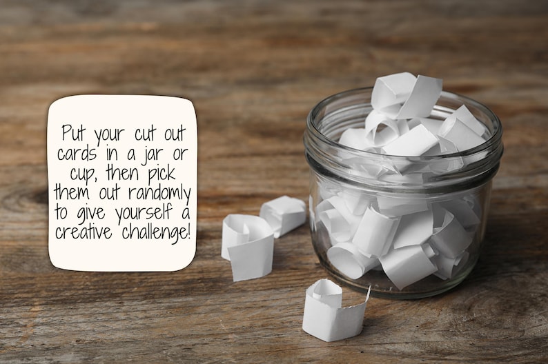 May include: A glass jar filled with white paper slips, with a few slips scattered on a wooden surface. The text on the white card reads: "Put your cut out cards in a jar or cup, then pick them out randomly to give yourself a creative challenge!"