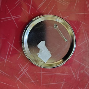 K&ouml;nnte beinhalten: Ein silberfarbener Ansteckbutton mit Sicherheitsnadel auf der R&uuml;ckseite. Der Button hat eine gl&auml;nzende, reflektierende Oberfl&auml;che. Ein St&uuml;ck wei&szlig;es Papier ist auf der R&uuml;ckseite befestigt. Der Button liegt auf einer roten Oberfl&auml;che mit wei&szlig;en Linien.