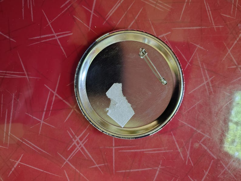 K&ouml;nnte beinhalten: Ein silberfarbener Ansteckbutton mit Sicherheitsnadel auf der R&uuml;ckseite. Der Button hat eine gl&auml;nzende, reflektierende Oberfl&auml;che. Ein St&uuml;ck wei&szlig;es Papier ist auf der R&uuml;ckseite befestigt. Der Button liegt auf einer roten Oberfl&auml;che mit wei&szlig;en Linien.