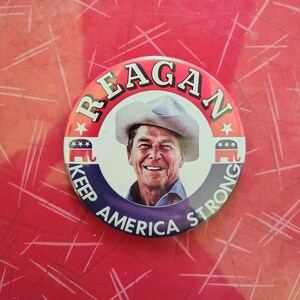 K&ouml;nnte beinhalten: Ein runder Button mit einem l&auml;chelnden Portr&auml;t von Ronald Reagan mit Cowboyhut. Der Button tr&auml;gt oben den Schriftzug "REAGAN" und unten "KEEP AMERICA STRONG". Der Button hat rote, wei&szlig;e und blaue Akzente.