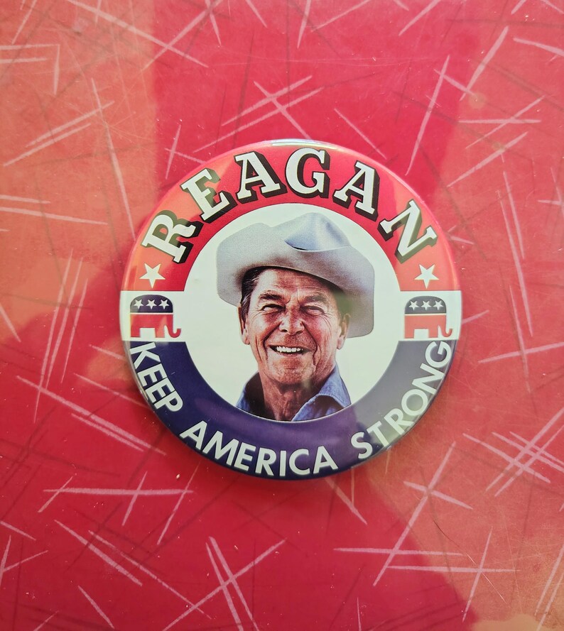 K&ouml;nnte beinhalten: Ein runder Button mit einem l&auml;chelnden Portr&auml;t von Ronald Reagan mit Cowboyhut. Der Button tr&auml;gt oben den Schriftzug "REAGAN" und unten "KEEP AMERICA STRONG". Der Button hat rote, wei&szlig;e und blaue Akzente.