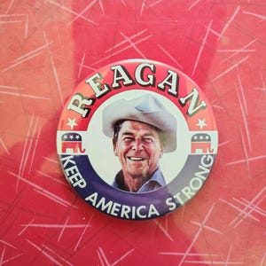 K&ouml;nnte beinhalten: Ein runder Button mit einem Portr&auml;t von Ronald Reagan mit Cowboyhut. Der Button hat den Text "REAGAN" oben und "KEEP AMERICA STRONG" unten. Der Hintergrund ist rot.
