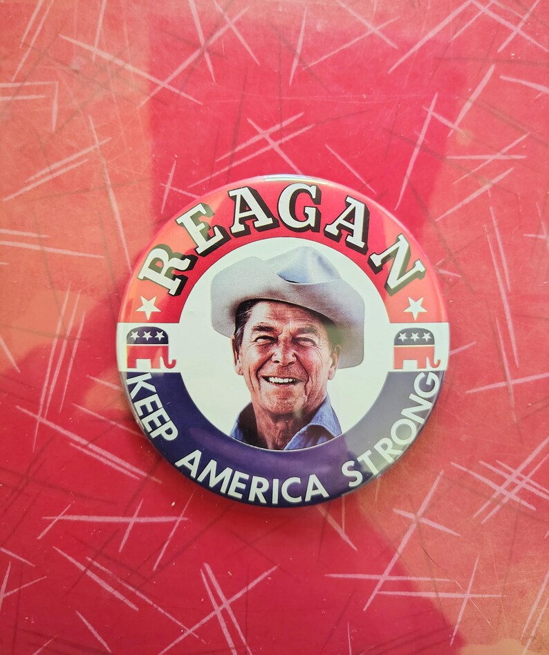 K&ouml;nnte beinhalten: Ein runder Button mit einem Portr&auml;t von Ronald Reagan mit Cowboyhut. Der Button hat den Text "REAGAN" oben und "KEEP AMERICA STRONG" unten. Der Hintergrund ist rot.