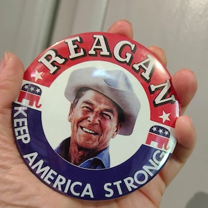 K&ouml;nnte beinhalten: Ein runder Button mit einem Portr&auml;t von Ronald Reagan mit Cowboyhut. Der Button hat ein rot-blaues Farbschema mit den Worten "REAGAN" und "KEEP AMERICA STRONG" aufgedruckt.