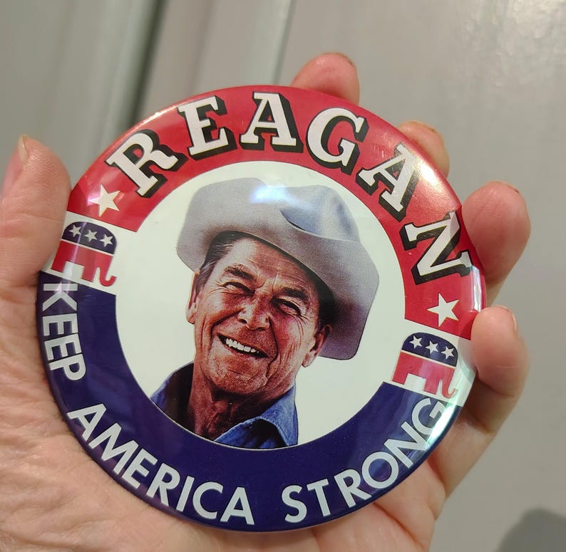 K&ouml;nnte beinhalten: Ein runder Button mit einem Portr&auml;t von Ronald Reagan mit Cowboyhut. Der Button hat ein rot-blaues Farbschema mit den Worten "REAGAN" und "KEEP AMERICA STRONG" aufgedruckt.