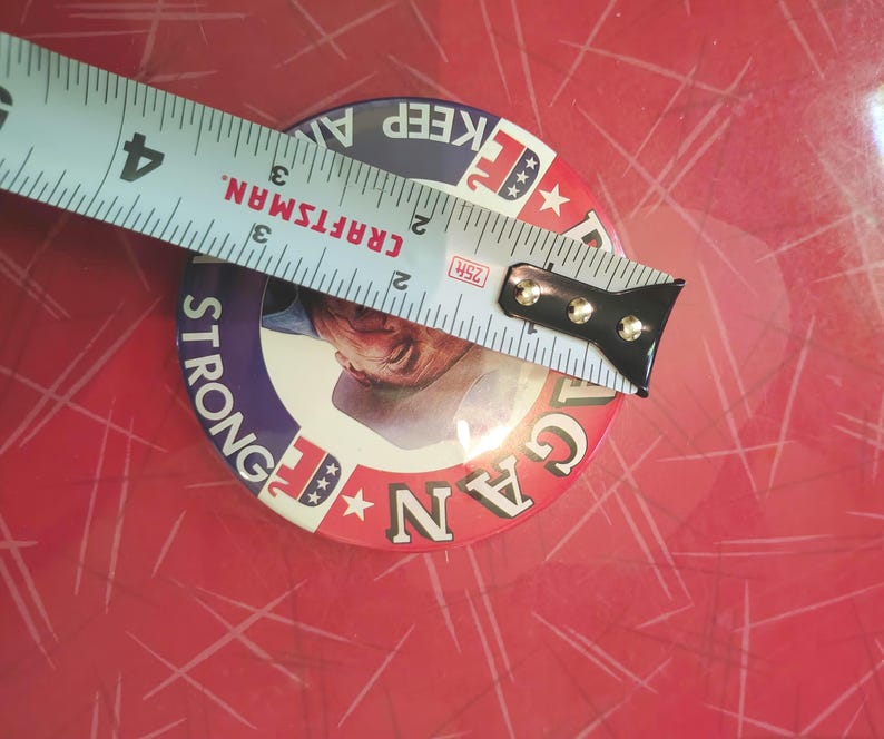 K&ouml;nnte beinhalten: Ein runder Button mit einem Portr&auml;t und den Worten "STRONG" und "REAGAN" in einem rot-wei&szlig;-blauen Design. Der Button hat einen Durchmesser von etwa 7,6 cm. Ein Craftsman-Ma&szlig;band liegt &uuml;ber dem Button.