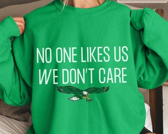 Może przedstawiać: Jaskrawozielona bluza z okrągłym dekoltem z białym napisem "NO ONE LIKES US WE DON'T CARE" wydrukowanym pogrubioną czcionką bezszeryfową. Pod tekstem znajduje się graficzna ilustracja zielono-białego orła trzymającego w szponach piłkę do futbolu.