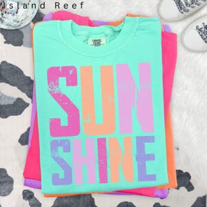 May include: A stack of colorful t-shirts. The top shirt is a light turquoise with the word "SUNSHINE" in large, distressed, multi-colored letters. The other shirts are pink, orange, and purple. The shirts are folded neatly on a white surface.