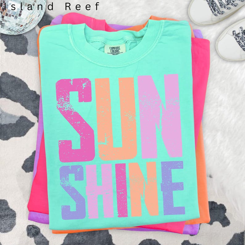 May include: A stack of colorful t-shirts. The top shirt is a light turquoise with the word "SUNSHINE" in large, distressed, multi-colored letters. The other shirts are pink, orange, and purple. The shirts are folded neatly on a white surface.