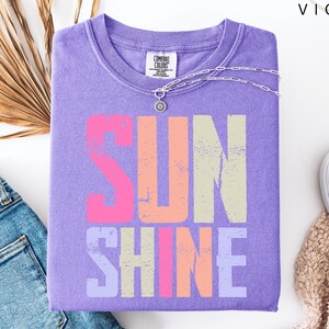 May include: A lilac-colored t-shirt with the word "SUNSHINE" printed in a distressed, multi-colored font. The shirt is folded and accessorized with a silver necklace. A pair of blue jeans and white sneakers are also in the image.