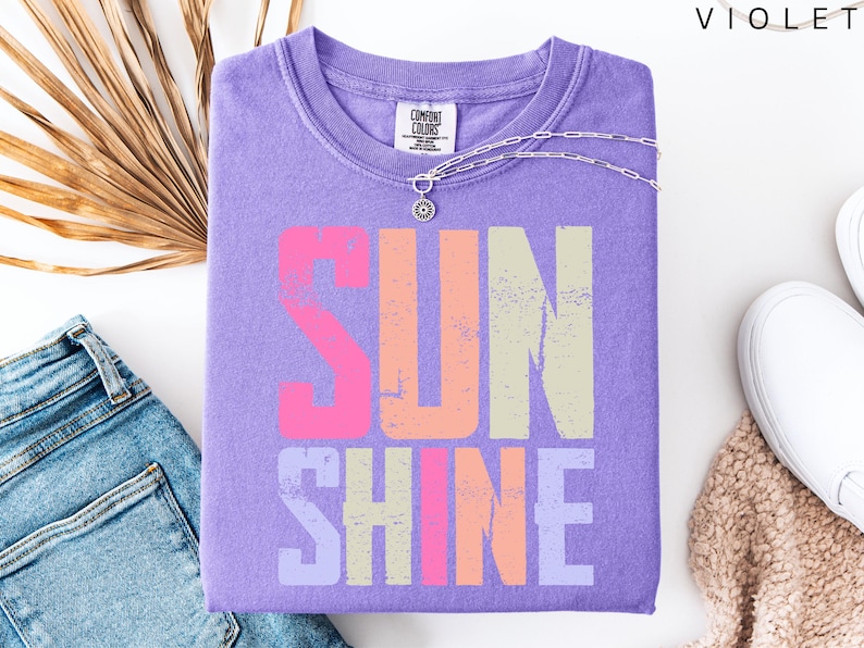 May include: A lilac-colored t-shirt with the word "SUNSHINE" printed in a distressed, multi-colored font. The shirt is folded and accessorized with a silver necklace. A pair of blue jeans and white sneakers are also in the image.