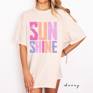 May include: Ivory-colored t-shirt with the word "SUNSHINE" in large, distressed, colorful letters. The word "SUN" is in pink, orange, and purple, while "SHINE" is in purple, pink, and orange. The shirt has short sleeves.