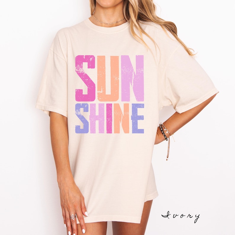 May include: Ivory-colored t-shirt with the word "SUNSHINE" in large, distressed, colorful letters. The word "SUN" is in pink, orange, and purple, while "SHINE" is in purple, pink, and orange. The shirt has short sleeves.