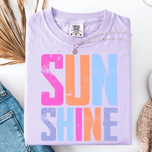 May include: A lavender Comfort Colors t-shirt with the word "SUNSHINE" in large, distressed, colorful letters. The letters are pink, orange, and blue. A silver necklace and white sneakers are also in the image.