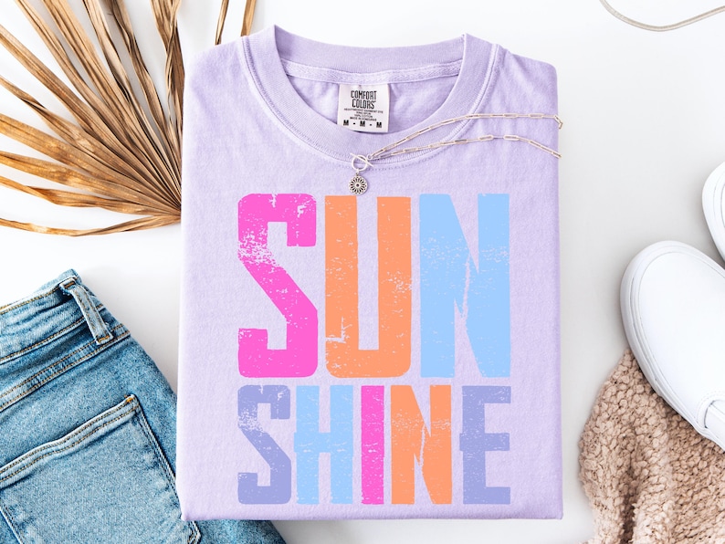 May include: A lavender Comfort Colors t-shirt with the word "SUNSHINE" in large, distressed, colorful letters. The letters are pink, orange, and blue. A silver necklace and white sneakers are also in the image.