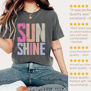 May include: A dark gray t-shirt with the word "SUNSHINE" in a colorful, distressed font. The shirt is worn with blue jeans. Customer reviews are displayed on the right side of the image.