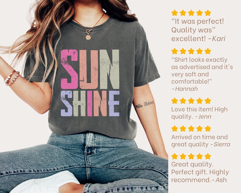 May include: A dark gray t-shirt with the word "SUNSHINE" in a colorful, distressed font. The shirt is worn with blue jeans. Customer reviews are displayed on the right side of the image.