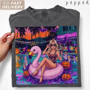 Op de afbeelding: Een antracietgrijs t-shirt met een afbeelding van een persoon met blond haar en een zonnebril op een roze flamingo zwemband. De scène bevat een zwembad, pompoenen, paarse vleermuizen en een neonbord met de tekst Summerween.