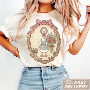 Puede incluir: Camiseta color crema con una ilustración de estilo vintage de un ganso con gorro y delantal, sosteniendo una cesta de flores. El diseño está enmarcado por un óvalo decorativo con un lazo rosa en la parte superior. El delantal dice "The Cottage Florist".