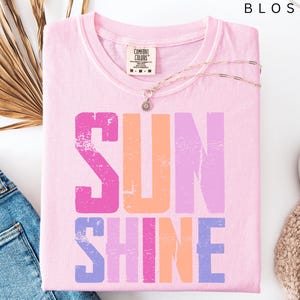 May include: A light pink Comfort Colors t-shirt with the word "SUNSHINE" printed in distressed pink, orange, and purple. A gold necklace with a pendant rests on the shirt. The shirt is folded next to blue jeans, a beige sweater, and white sneakers.