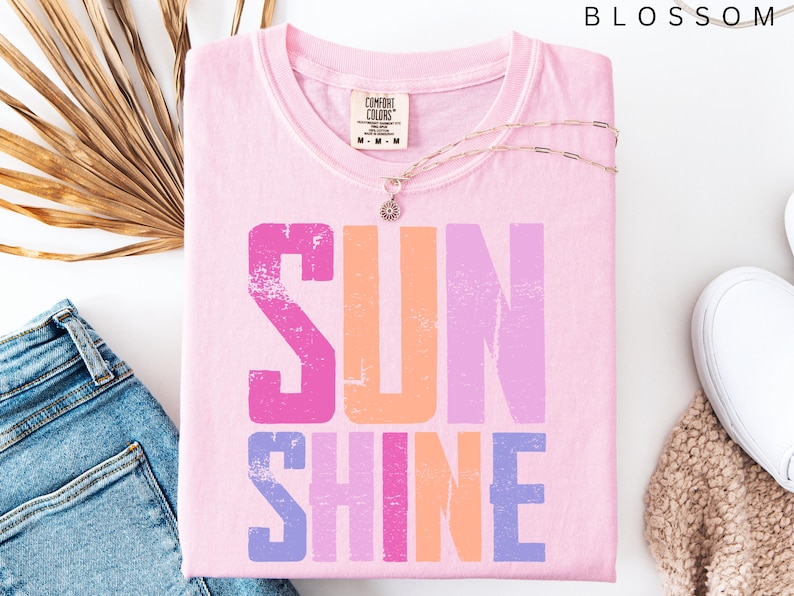 May include: A light pink Comfort Colors t-shirt with the word "SUNSHINE" printed in distressed pink, orange, and purple. A gold necklace with a pendant rests on the shirt. The shirt is folded next to blue jeans, a beige sweater, and white sneakers.