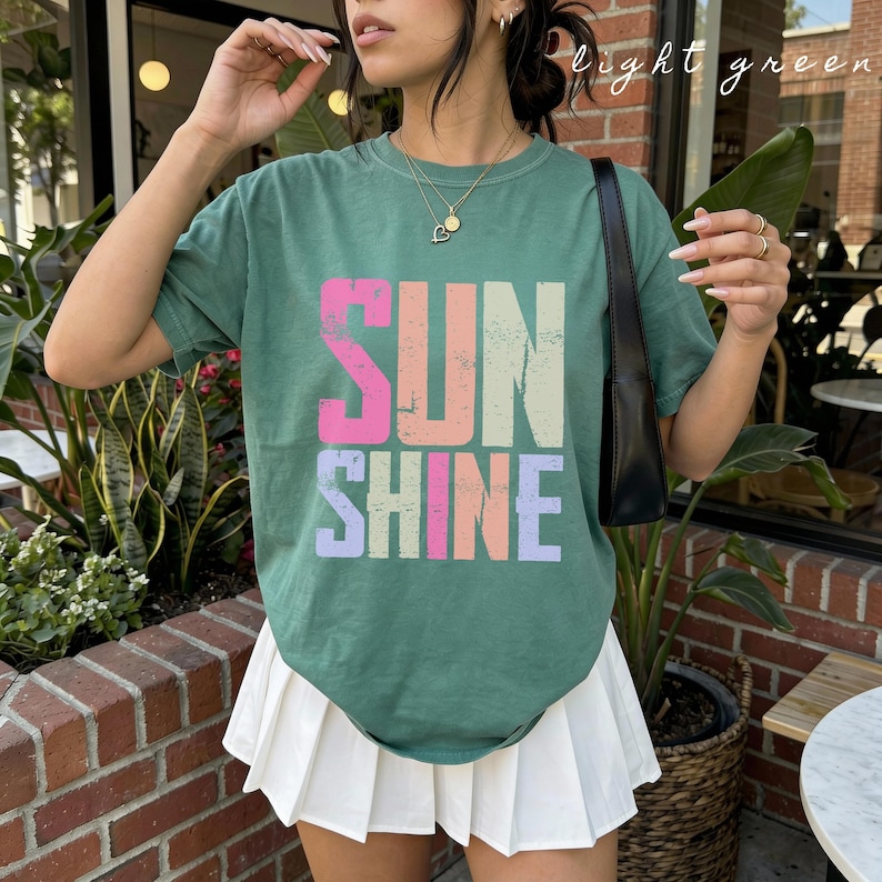 May include: A light green t-shirt with the word "SUNSHINE" in large, colorful letters. The text is in pink, orange, light green, and purple. The shirt is paired with a white pleated skirt.