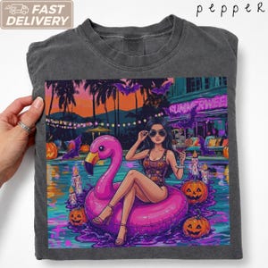 Op de afbeelding: Een antracietgrijs t-shirt met een grafische print van een persoon met een zonnebril en badpak, zittend op een roze flamingo zwemband in een zwembad bij nacht. De scène bevat brandende kaarsen, pompoenen, vleermuizen en palmbomen.
