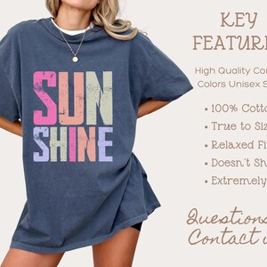 May include: A navy blue unisex t-shirt with the word "SUNSHINE" in colorful, distressed lettering. The shirt features a relaxed fit and is made of 100% cotton. Additional text on the right side lists key features of the shirt.