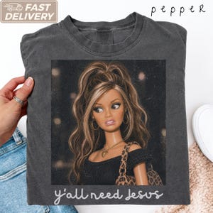 May include: A charcoal gray crew neck t-shirt featuring a graphic print of a doll with long brown hair, gold hoop earrings, and a gold heart necklace. The doll wears a black top and a leopard print bag. The text "y'all need jesus" is printed below.