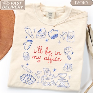 May include: An ivory t-shirt featuring the red text i'll be in my office surrounded by blue line art illustrations of baking items including a piping bag, milk carton, chef hat, flour bag, oven mitt, cupcake, mixer, rolling pin, eggs, apron, and scale.