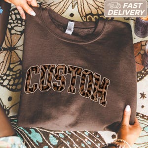 Könnte beinhalten: Ein dunkelbraunes Sweatshirt mit Rundhalsausschnitt und dem Wort CUSTOM in einer großen, geschwungenen College-Schriftart. Die Buchstaben haben ein braun-schwarzes Leopardenmuster mit einer dünnen weißen Umrandung und einem Rippkragen.