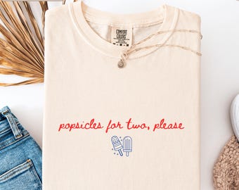 Puede incluir: Una camiseta de color marfil con el texto en rojo "popsicles for two, please" sobre un gráfico de líneas azules de dos polos. La camiseta tiene cuello redondo y una etiqueta de la marca Comfort Colors en el interior, con un collar dorado sobre el cuello.